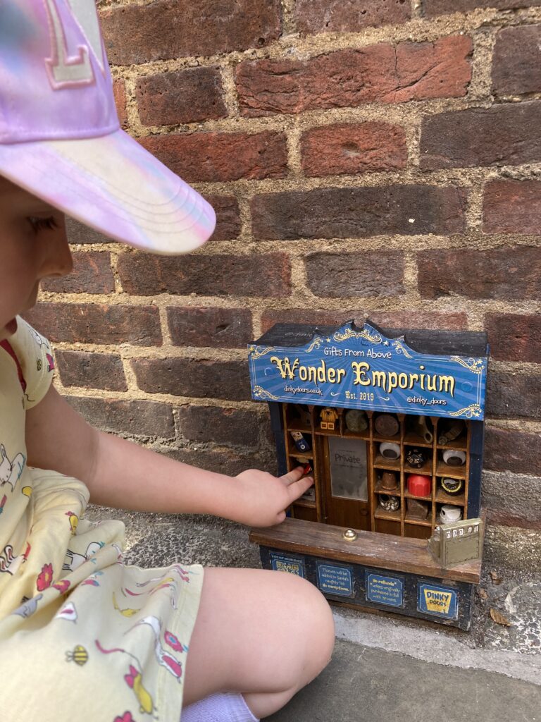 Young girl in a pink cap peering into the Wonder Emporium dinky door on a Cambridge brick wall.