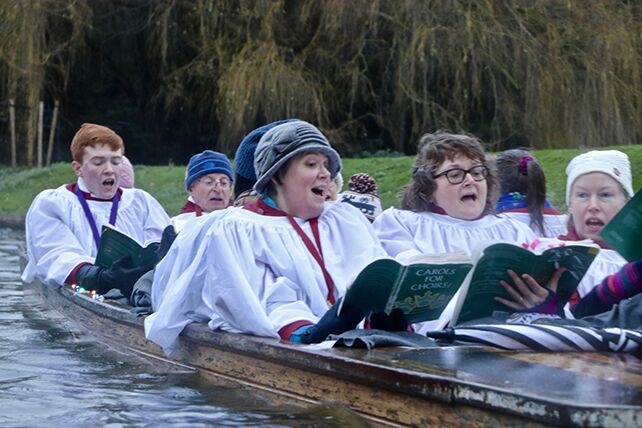 Choir members in white robes singing from carol books aboard a punt on the River Cam.