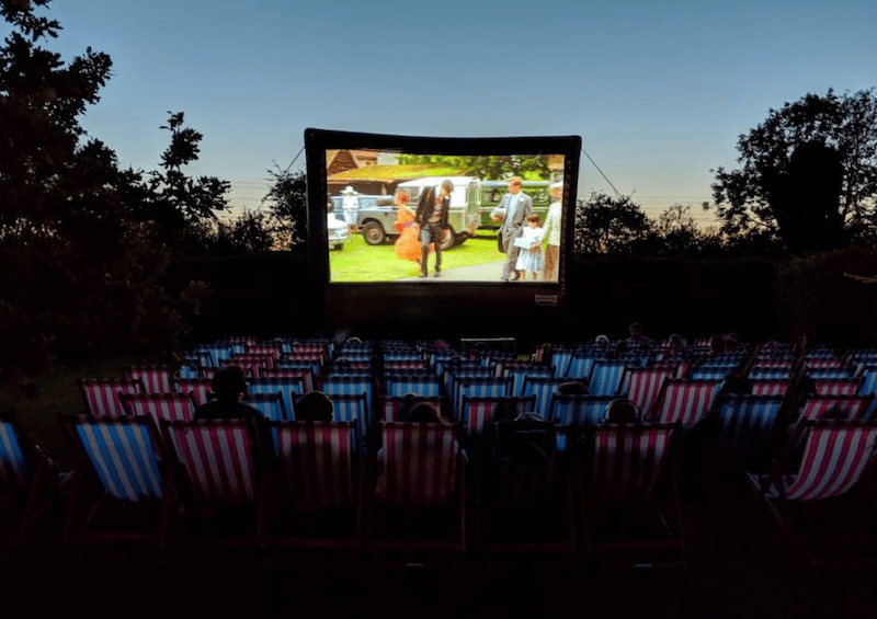 Audience in striped deckchairs watching a film on a large outdoor cinema screen at dusk.