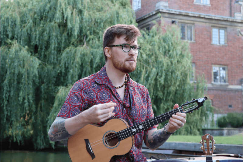 Bearded musician playing a ukulele on a punt by the River Cam during Folk on a Boat.