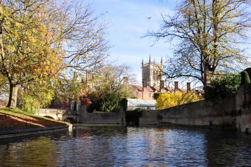 Autumn trees reflected on the River Cam with a college tower rising beyond the riverbank.