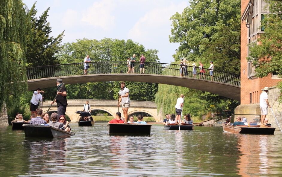 Busy summer afternoon with punts passing under a footbridge on the River Cam.