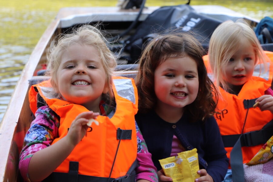 Three grinning young girls in orange life jackets enjoying a family punting trip in Cambridge.