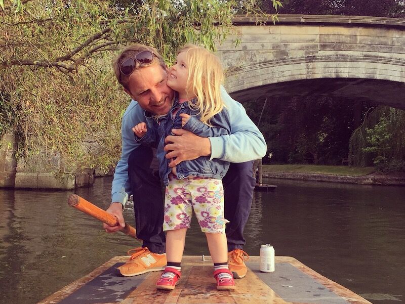 Young girl kissing her dad's cheek as he holds her on the back of a punt near a stone bridge.