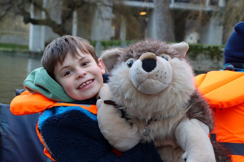 Smiling boy in a life jacket hugging a large toy wolf during a family punting trip in Cambridge.