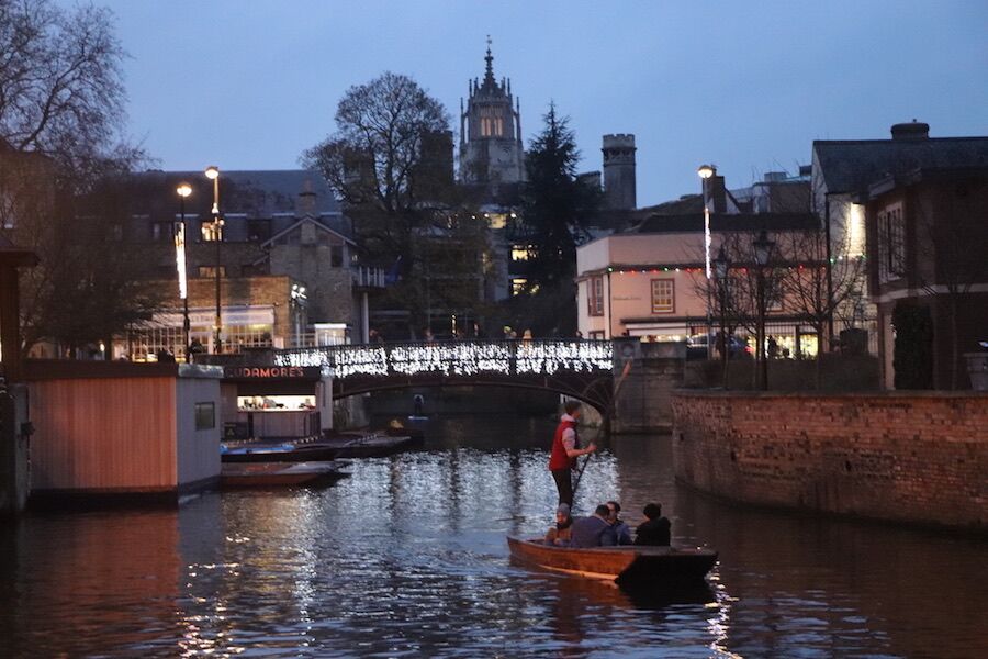 Lone punt on the river at dusk with festive lights and Cambridge rooftops behind.
