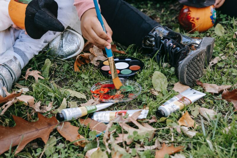 Child painting with a face paint palette on grass scattered with autumn leaves.