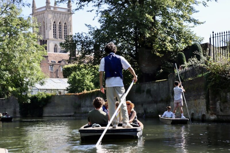 Chauffeur punting a boat of customers along the River Cam with a church tower in view.