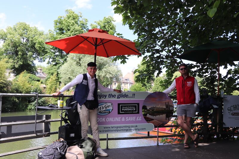 Tom and Josh standing by the Let's Go Punting cashier station under a red parasol by the river.