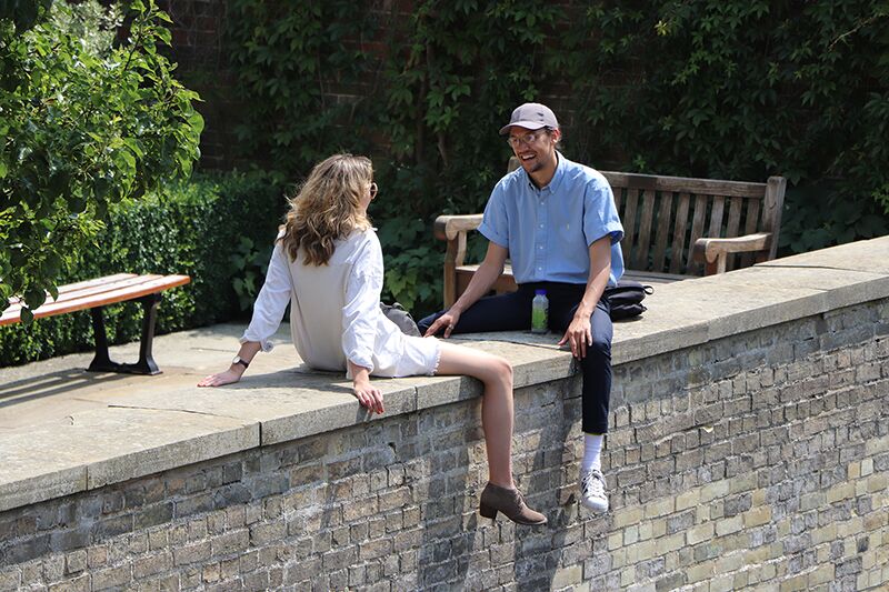 Two people sitting on the brick riverside wall at Trinity Hall beside a bench